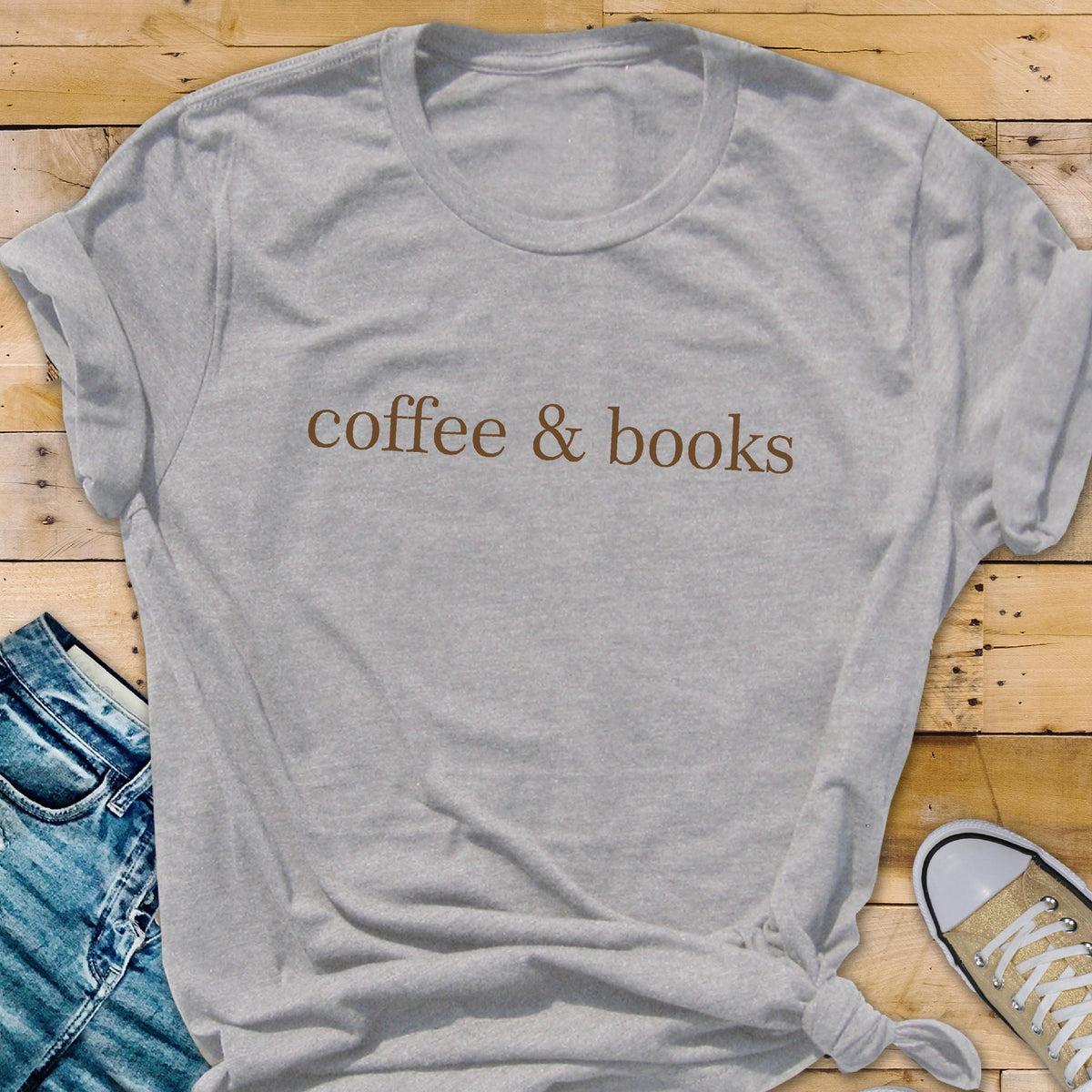 May include: A light gray short-sleeved t-shirt featuring the text "coffee & books" printed in a brown serif font across the chest. The garment has a crew neckline and a standard fit, showcasing a simple design with clear, legible lettering.