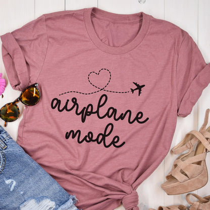 May include: Dusty rose t-shirt with the words "airplane mode" in a cursive font, accompanied by a heart and airplane graphic. The shirt is styled with sunglasses, denim shorts, and platform sandals.