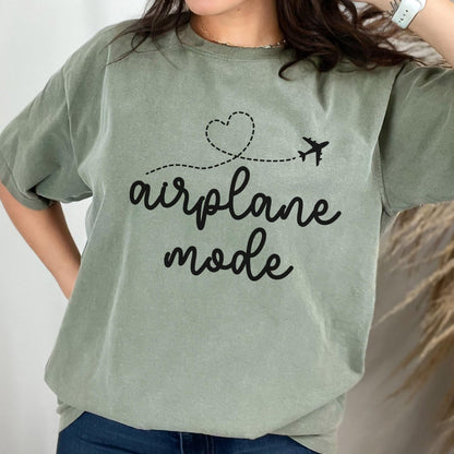 May include: Sage green t-shirt with the words "airplane mode" in black script. A dotted line with a heart and airplane graphic is above the text. The shirt has short sleeves and a relaxed fit.