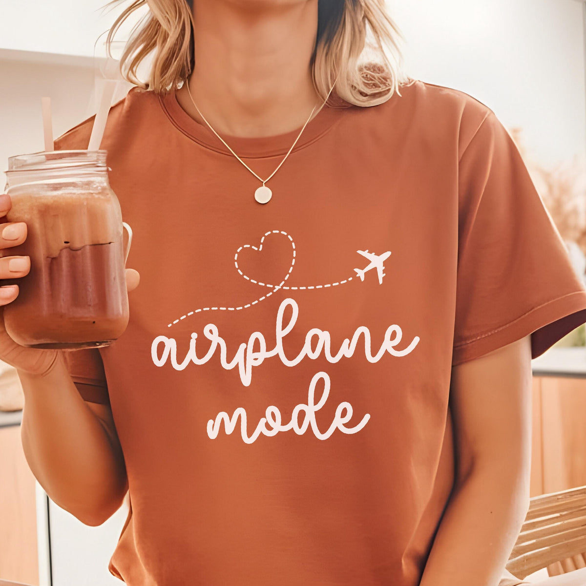 May include: A rust-colored t-shirt with the words "airplane mode" in white script. A white airplane graphic follows a dotted heart shape. The person is wearing a gold necklace and holding a glass of a brown beverage.