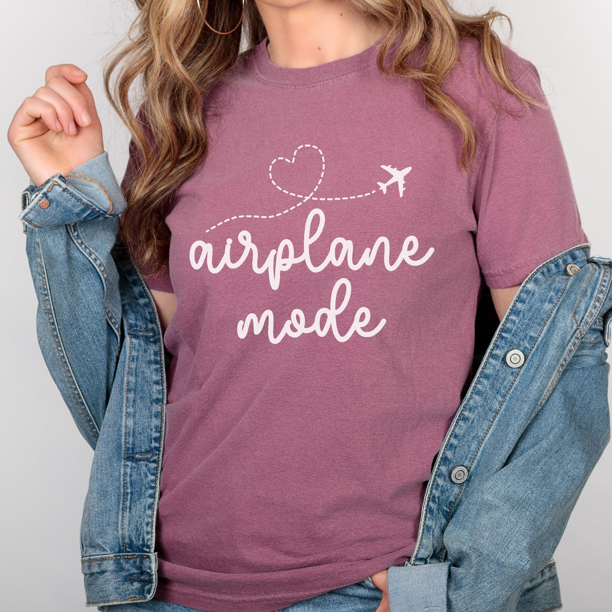 May include: Mauve t-shirt with the words "airplane mode" in white script. A white airplane graphic and a heart-shaped dotted line are above the text. The person is wearing a denim jacket.