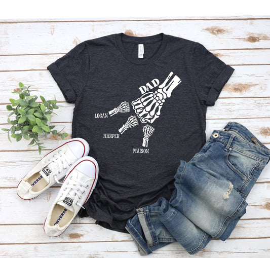 May include: A dark grey t-shirt with a white skeleton hand graphic. The hand is holding three smaller skeleton hands, each labeled with a name: Logan, Harper, and Maison. The text "DAD" is above the large hand.