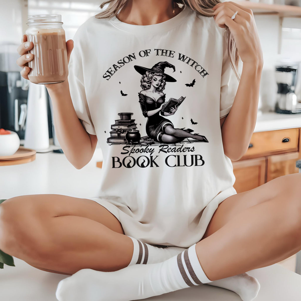 May include: Cream-colored t-shirt featuring a black and white illustration of a witch reading a book, with the text "Season of the Witch" and "Spooky Readers Book Club." The shirt is worn with white socks and a drink in a glass jar.
