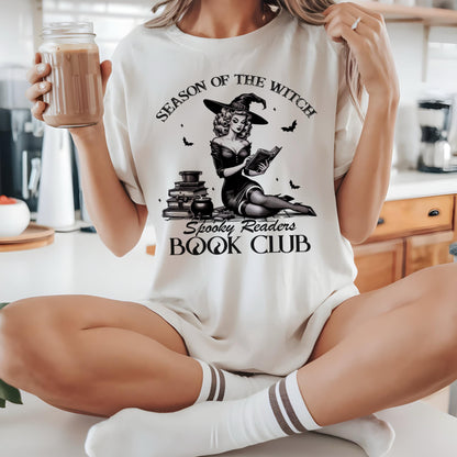 May include: Cream-colored t-shirt featuring a black and white illustration of a witch reading a book, with the text "Season of the Witch" and "Spooky Readers Book Club." The shirt is worn with white socks and a drink in a glass jar.
