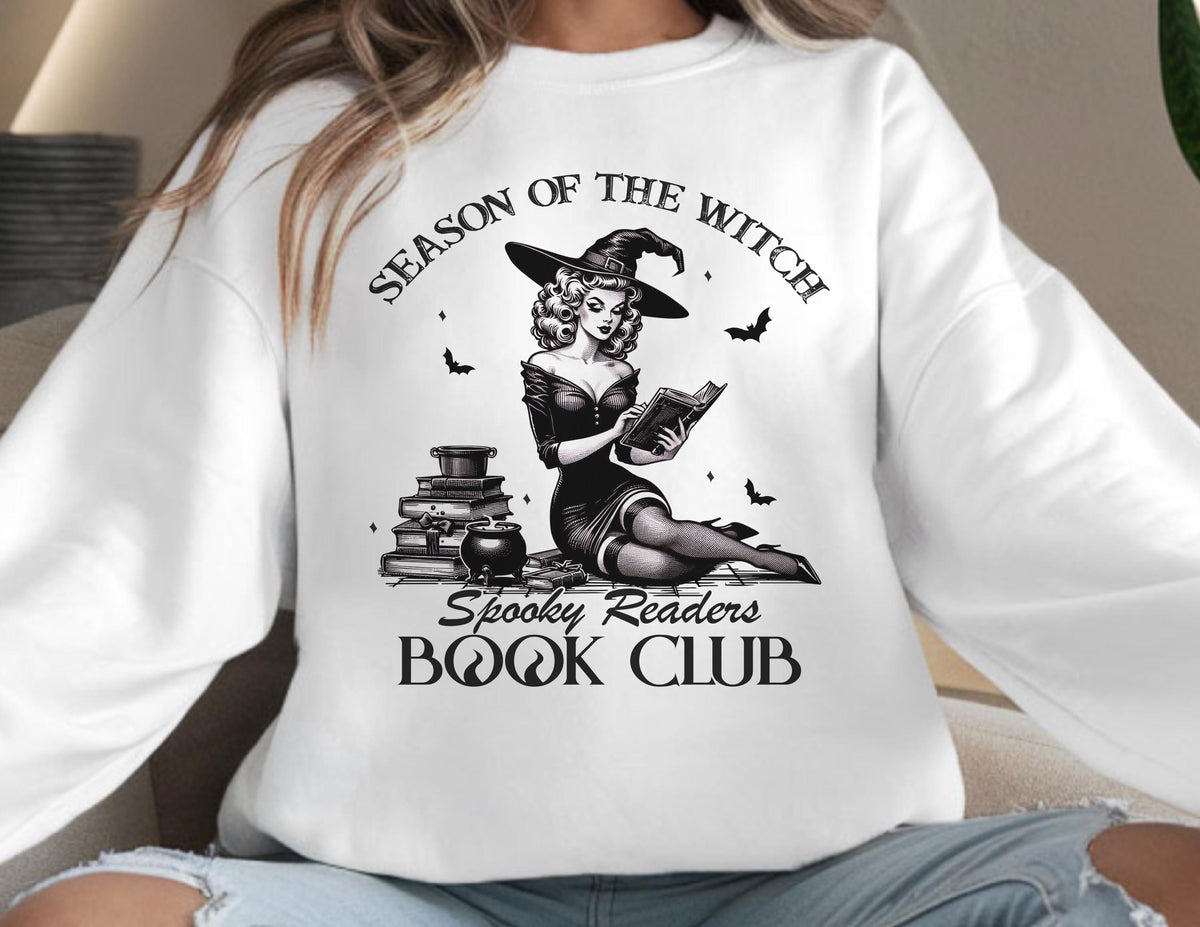 May include: White sweatshirt featuring a black and white illustration of a witch reading a book. The design includes the text "Season of the Witch" and "Spooky Readers Book Club", along with bats and a cauldron.