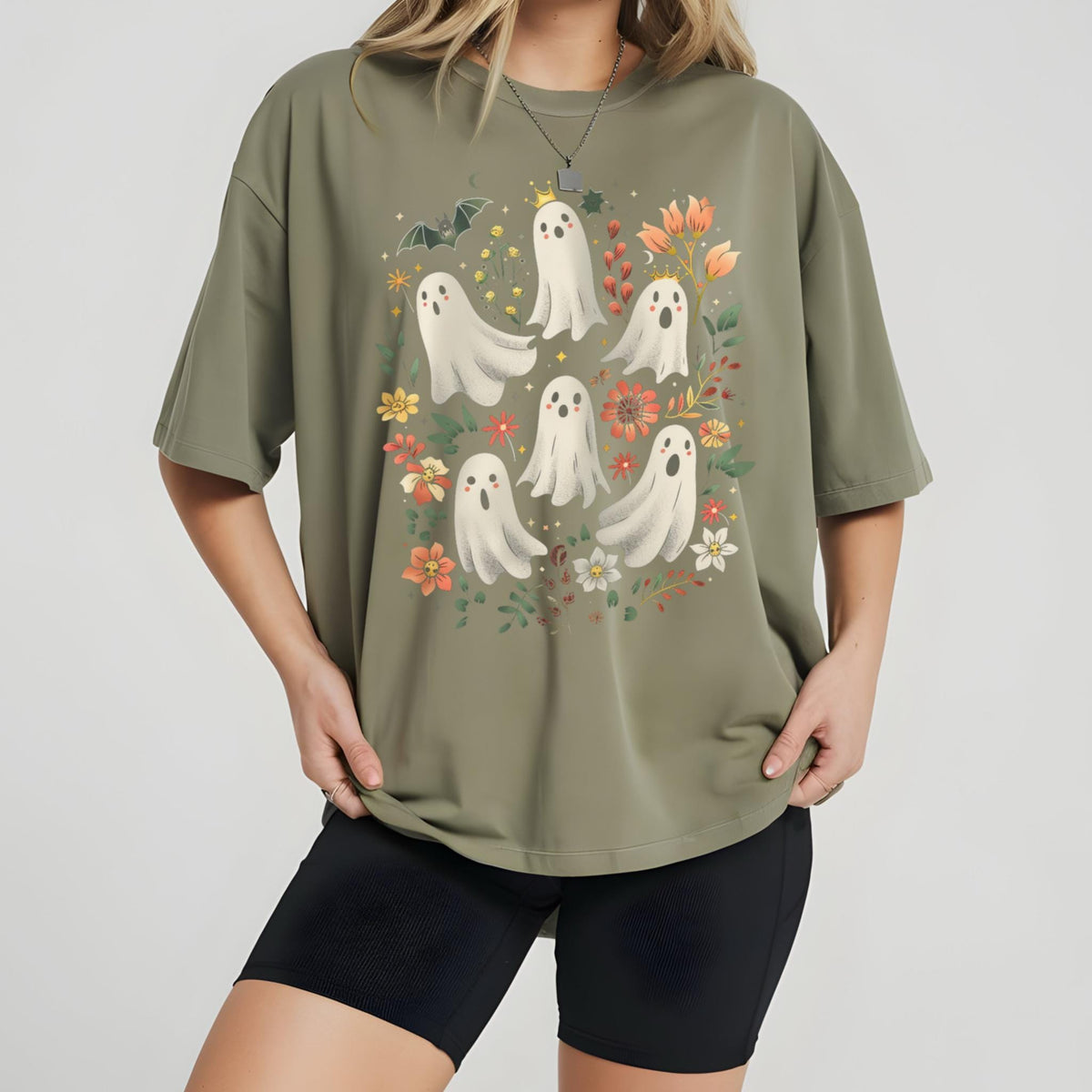 May include: Olive green t-shirt featuring a whimsical design of white ghosts with floral accents and a bat. The ghosts have small crowns and are surrounded by colorful flowers. A fun and festive graphic tee for Halloween.