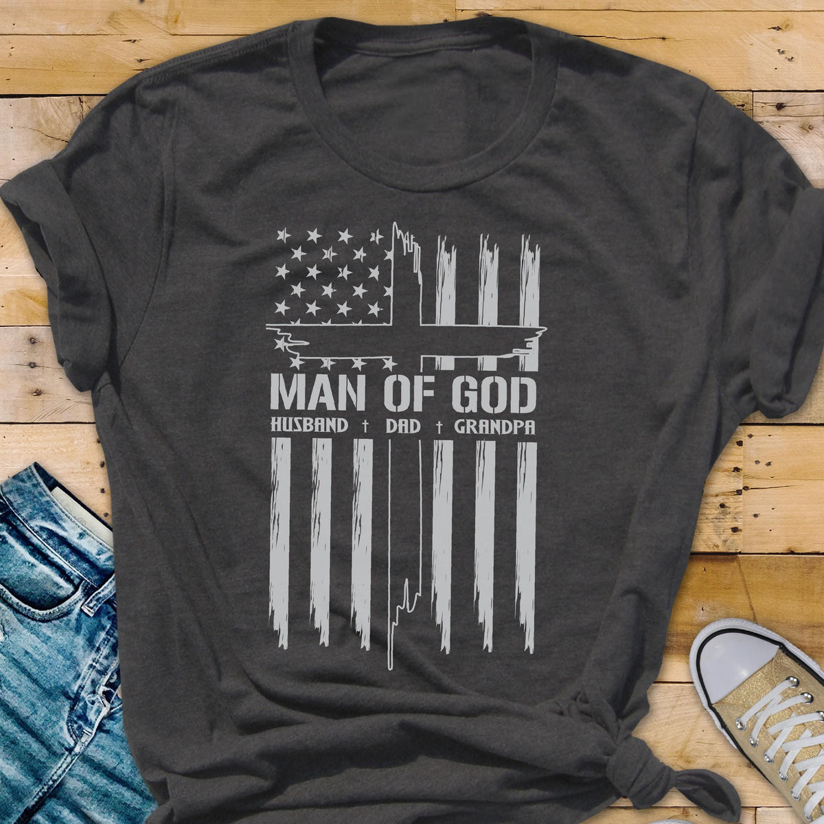May include: Dark gray t-shirt featuring a white graphic of an American flag with a cross design. The text reads "MAN OF GOD" above "HUSBAND + DAD + GRANDPA". The shirt is made of a soft material.