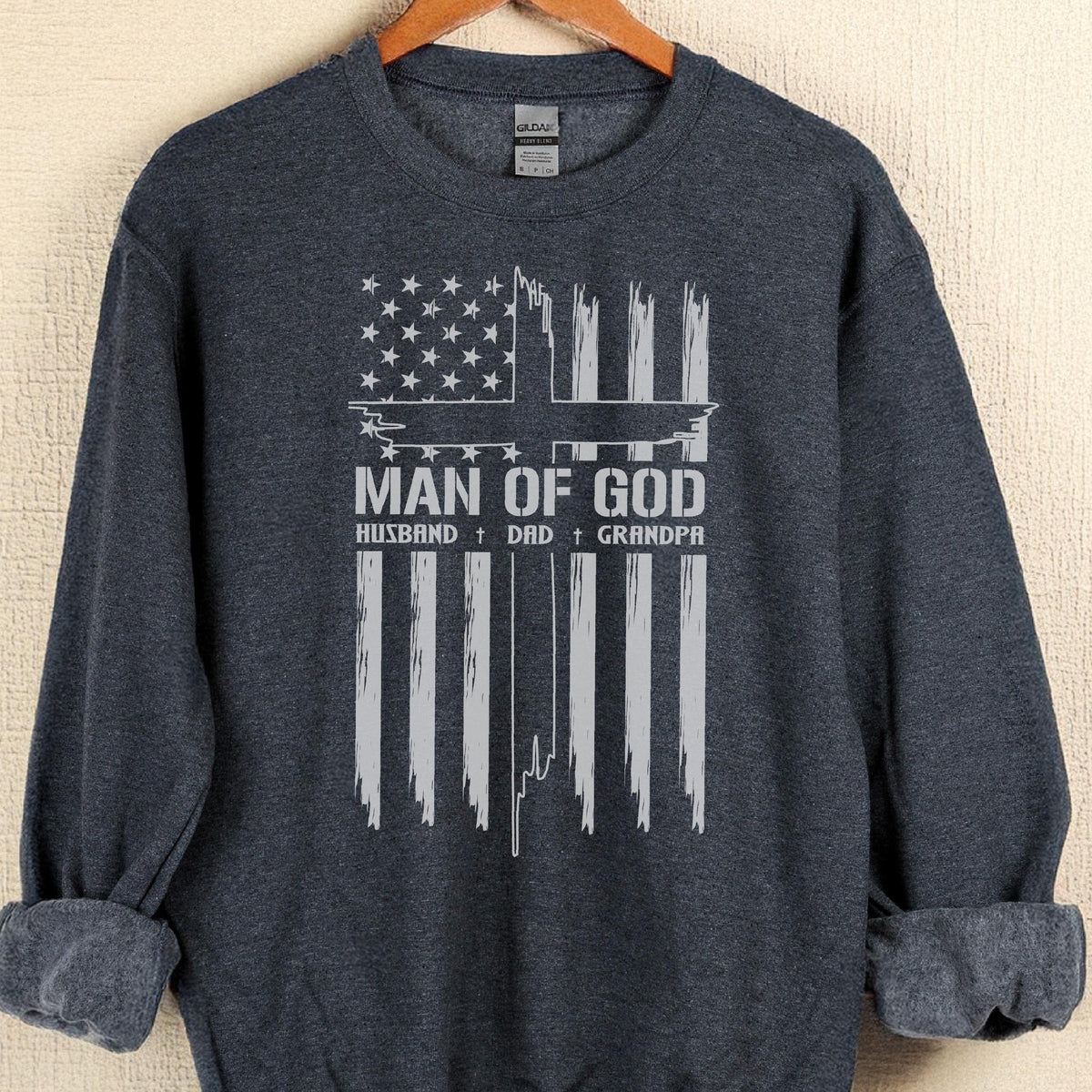 May include: Dark navy blue sweatshirt with a distressed white graphic. The graphic features an American flag design with the text "MAN OF GOD" and the words "HUSBAND + DAD + GRANDPA" below it.