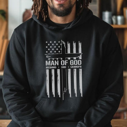 May include: Black hoodie with a white graphic design. The design features an American flag motif with a cross and the words "MAN OF GOD," "HUSBAND," "DAD," and "GRANDPA" in a bold, readable font. The hoodie has a drawstring hood and a front pocket.