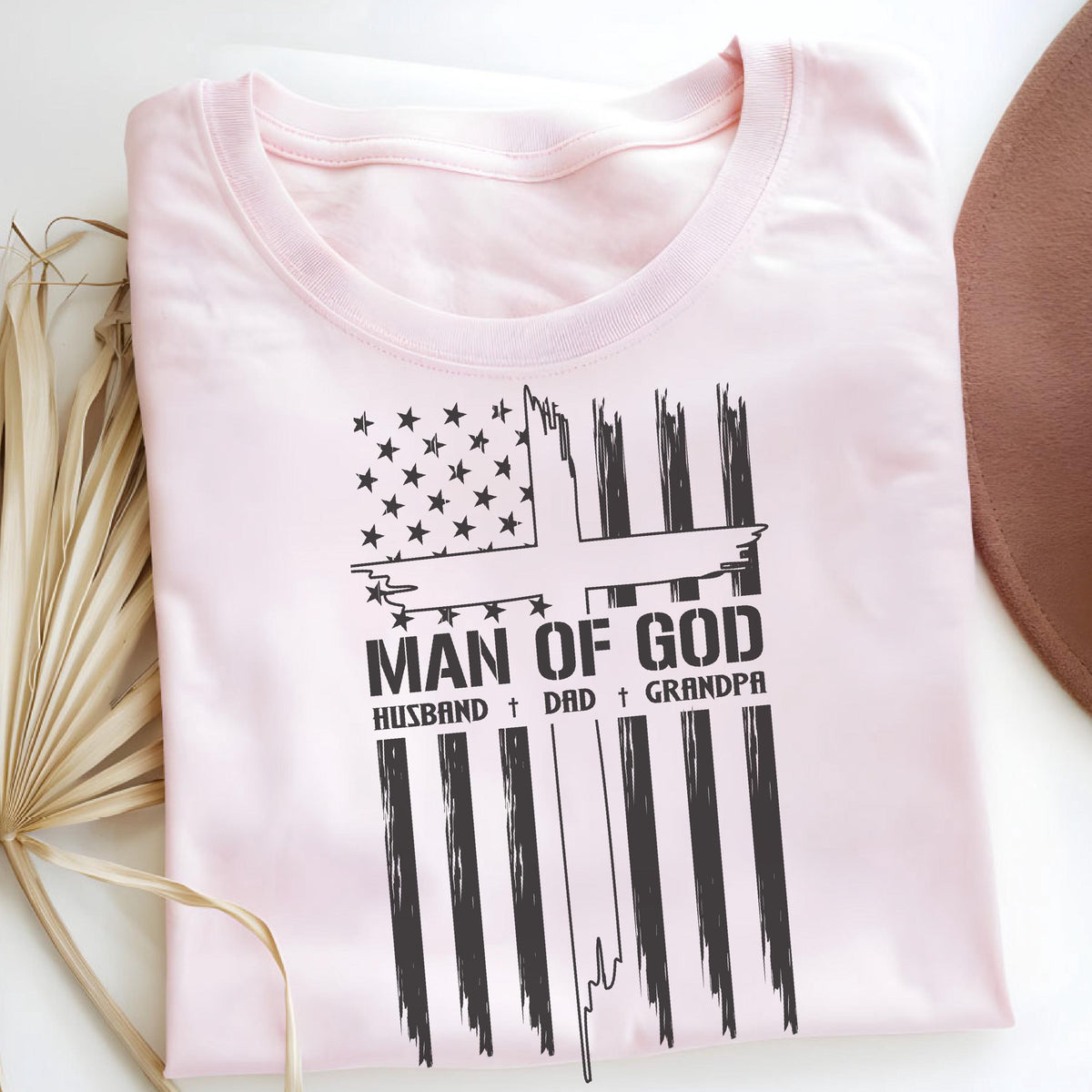 May include: A light pink t-shirt with a black graphic design. The design features a cross and the text "MAN OF GOD" above "HUSBAND + DAD + GRANDPA". The shirt is folded on a white surface.