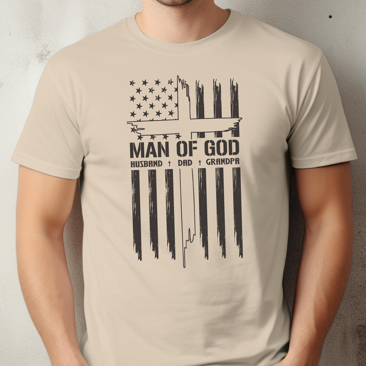 May include: Beige t-shirt featuring a distressed American flag design with a cross and the text "MAN OF GOD." Below the text are the words "HUSBAND + DAD + GRANDPA" in a simple font. The shirt is a neutral color.