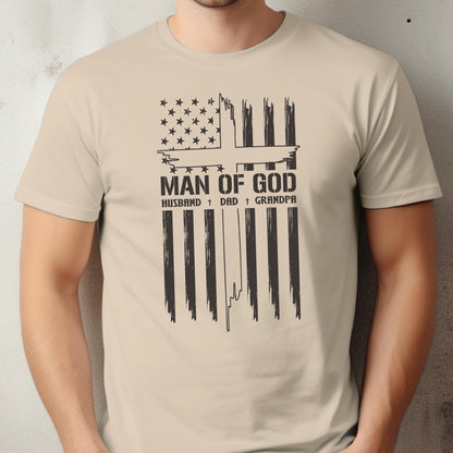 May include: Beige t-shirt featuring a distressed American flag design with a cross and the text "MAN OF GOD." Below the text are the words "HUSBAND + DAD + GRANDPA" in a simple font. The shirt is a neutral color.