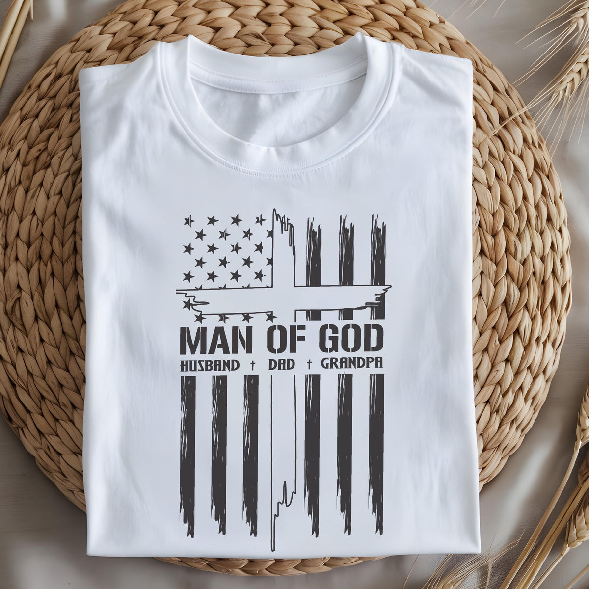 May include: White t-shirt with a graphic design featuring a cross and the text "MAN OF GOD, HUSBAND, DAD, GRANDPA". The design incorporates elements of the American flag. The shirt is displayed on a woven surface.
