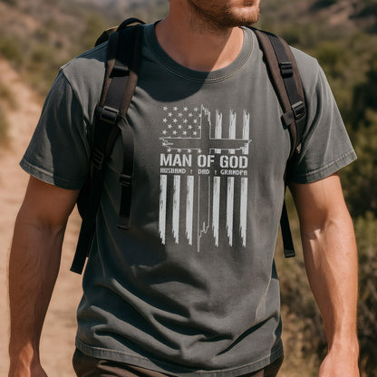 May include: Dark gray t-shirt with a white graphic of the American flag and a cross design. The text "MAN OF GOD" is above the words "HUSBAND † DAD † GRANDPA". The shirt is made of a soft material.