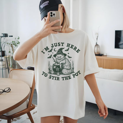 May include: Cream-colored t-shirt with a graphic of a cat wearing a witch hat, stirring a cauldron. The text reads "I'M JUST HERE TO STIR THE POT" in green. The design includes bats, candles, and stars, creating a Halloween theme.