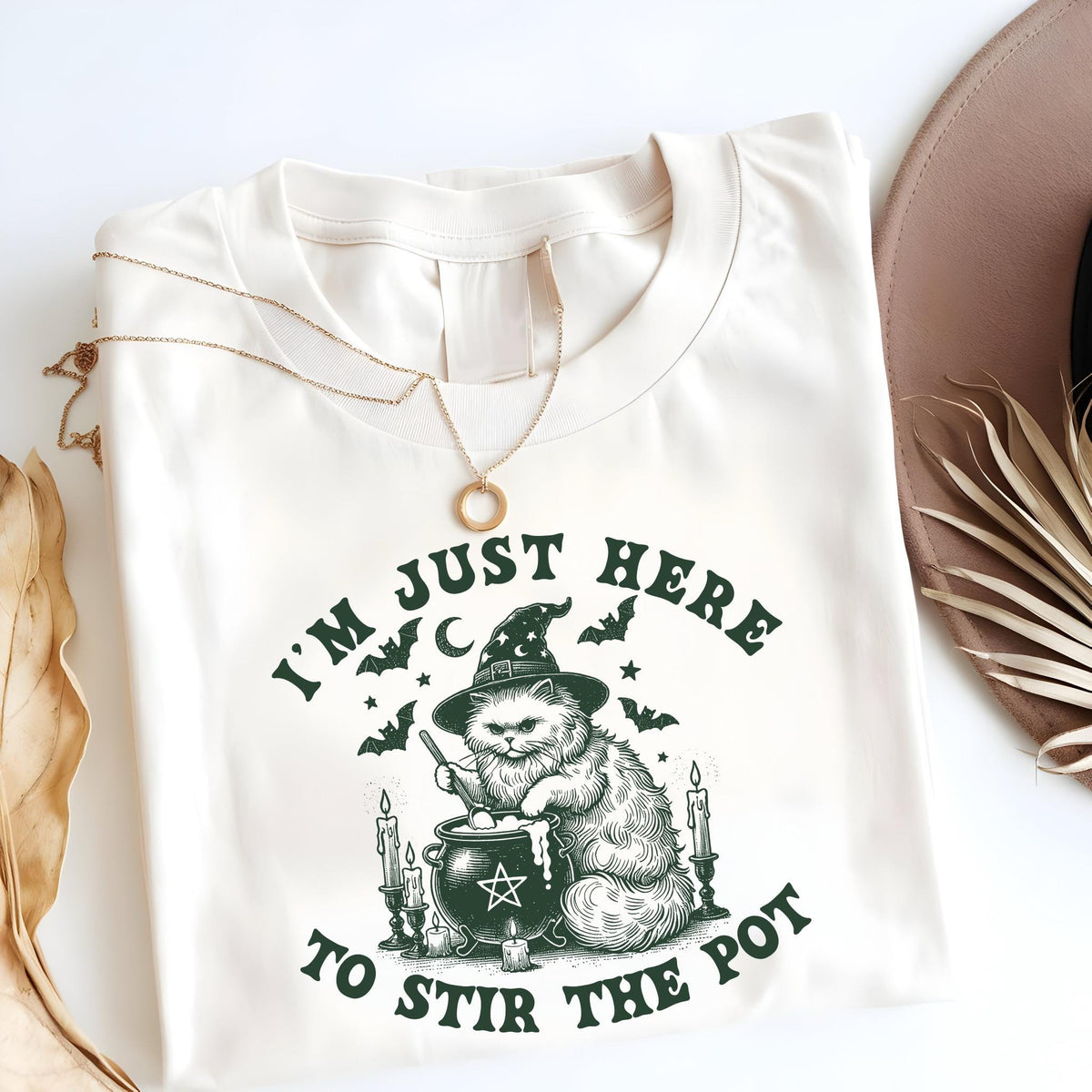 May include: Cream-colored t-shirt featuring a whimsical illustration of a cat wearing a witch's hat, stirring a cauldron. The text "I'M JUST HERE TO STIR THE POT" is printed in dark green. A gold necklace is draped on the shirt.