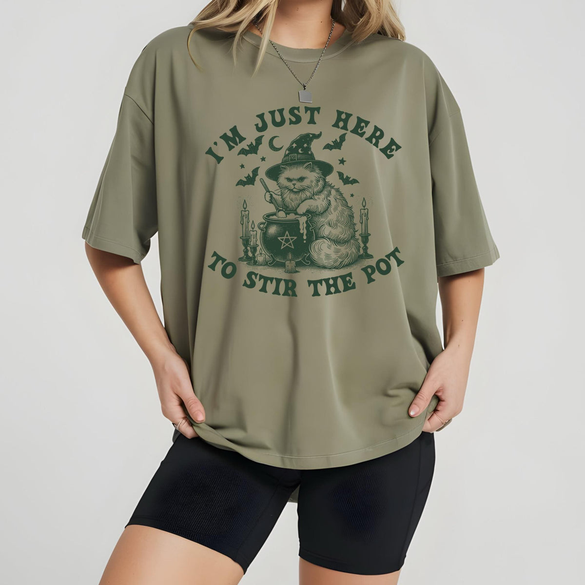 May include: Olive green t-shirt with a graphic of a cat wearing a witch hat, stirring a cauldron. The text reads "I'M JUST HERE TO STIR THE POT" in dark green. The shirt is paired with black shorts.
