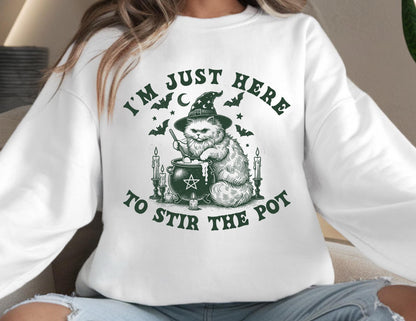 May include: White sweatshirt with a green graphic of a cat wearing a witch hat, stirring a cauldron. The text reads "I'M JUST HERE TO STIR THE POT" in green. Bats, stars, and candles surround the cat and cauldron.