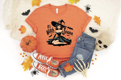 May include: Orange t-shirt with the text "It's Witch Season" and a graphic of a witch. The shirt is paired with blue jeans, orange sneakers, a pumpkin, and Halloween decorations.