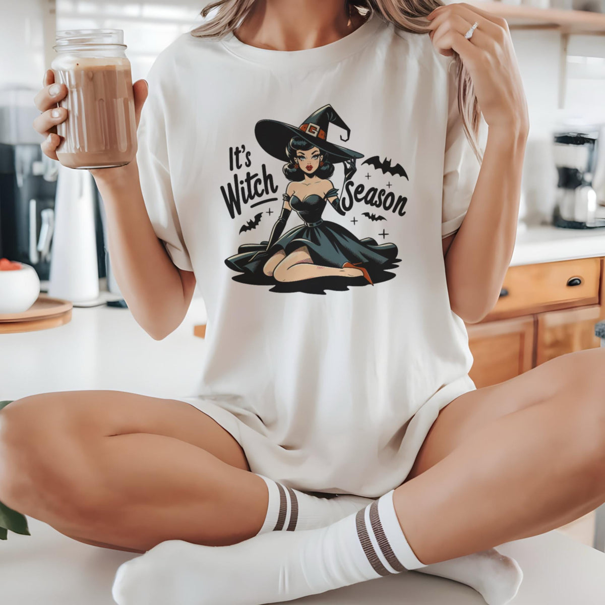 May include: Cream-colored t-shirt featuring a vintage-style illustration of a witch in a black dress and hat. The text "It's Witch Season" is above the witch. Bats and crosses are also included in the design. The shirt is a relaxed fit.