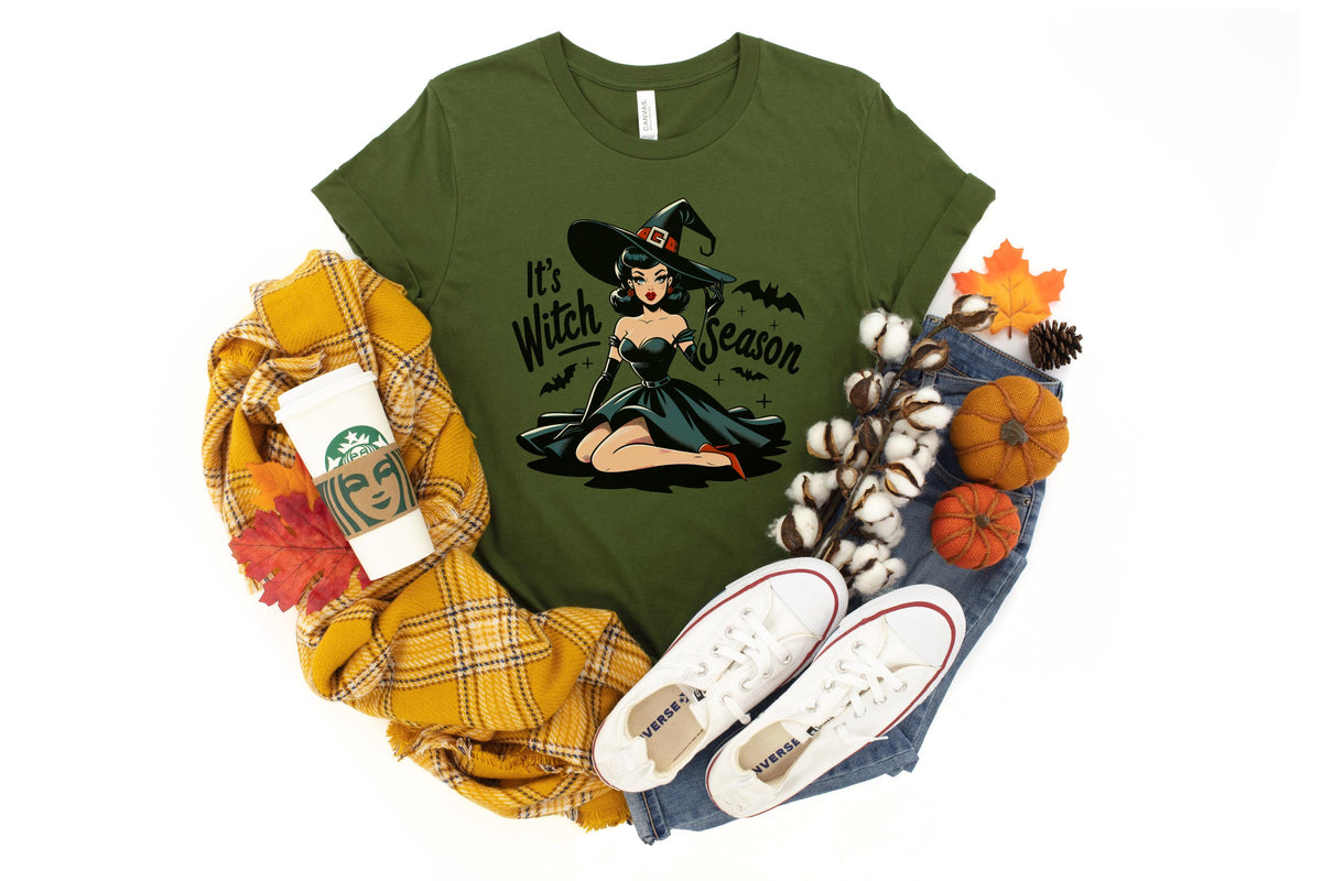 May include: Olive green t-shirt with the text "It's Witch Season" and a graphic of a witch. The shirt is styled with jeans, white sneakers, a yellow scarf, and fall-themed decorations.