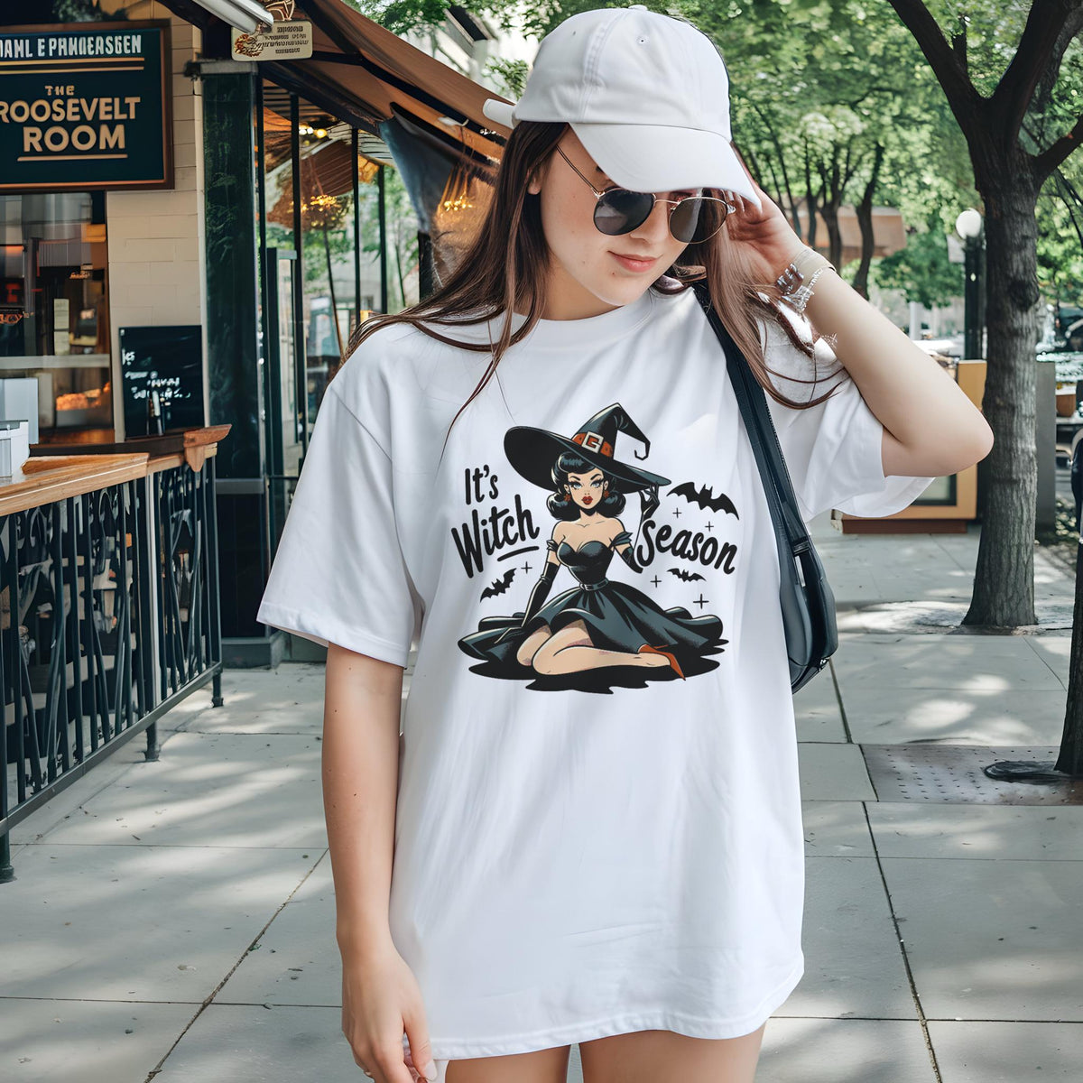 May include: White t-shirt featuring a graphic of a witch in a black dress and hat, with the text "It's Witch Season". The design includes bats and stars. A casual, oversized tee for Halloween.