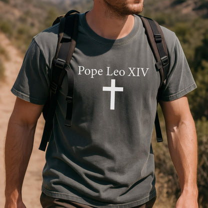 May include: Dark gray t-shirt with the text "Pope Leo XIV" above a white cross. The shirt is worn by a person with a backpack, set against a blurred outdoor background. The shirt's design is simple and features a religious symbol.