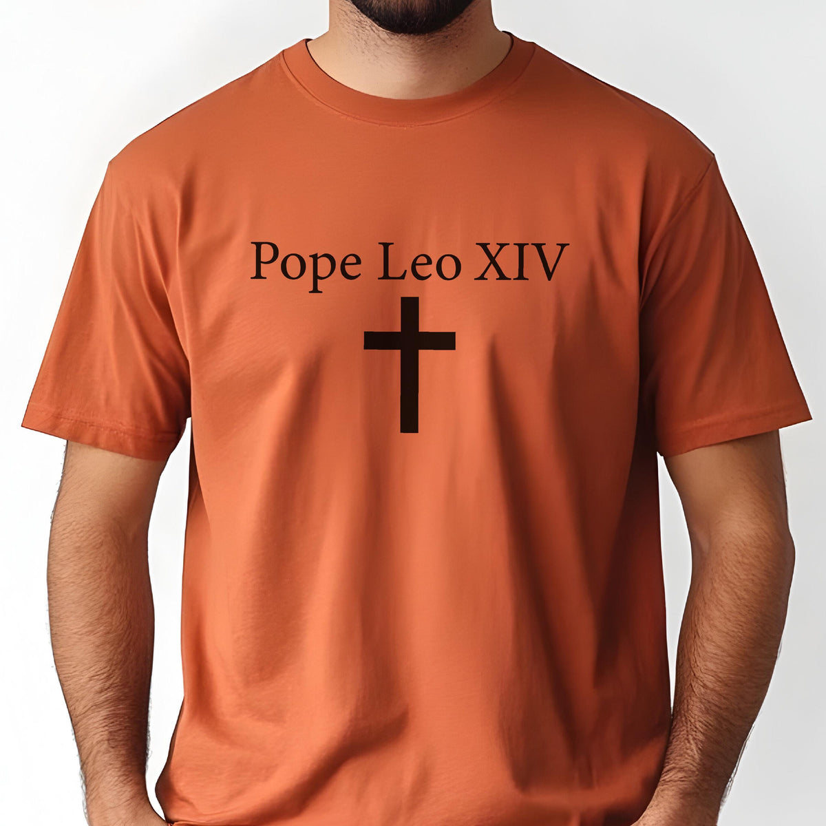 May include: A burnt orange t-shirt with the text "Pope Leo XIV" above a black cross. The short-sleeved shirt is made of a soft material and has a crew neck. The design is centered on the front of the shirt.