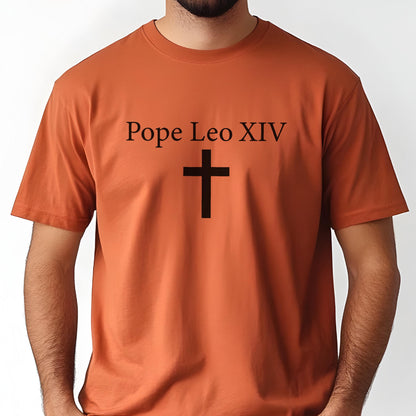 May include: A burnt orange t-shirt with the text "Pope Leo XIV" above a black cross. The short-sleeved shirt is made of a soft material and has a crew neck. The design is centered on the front of the shirt.