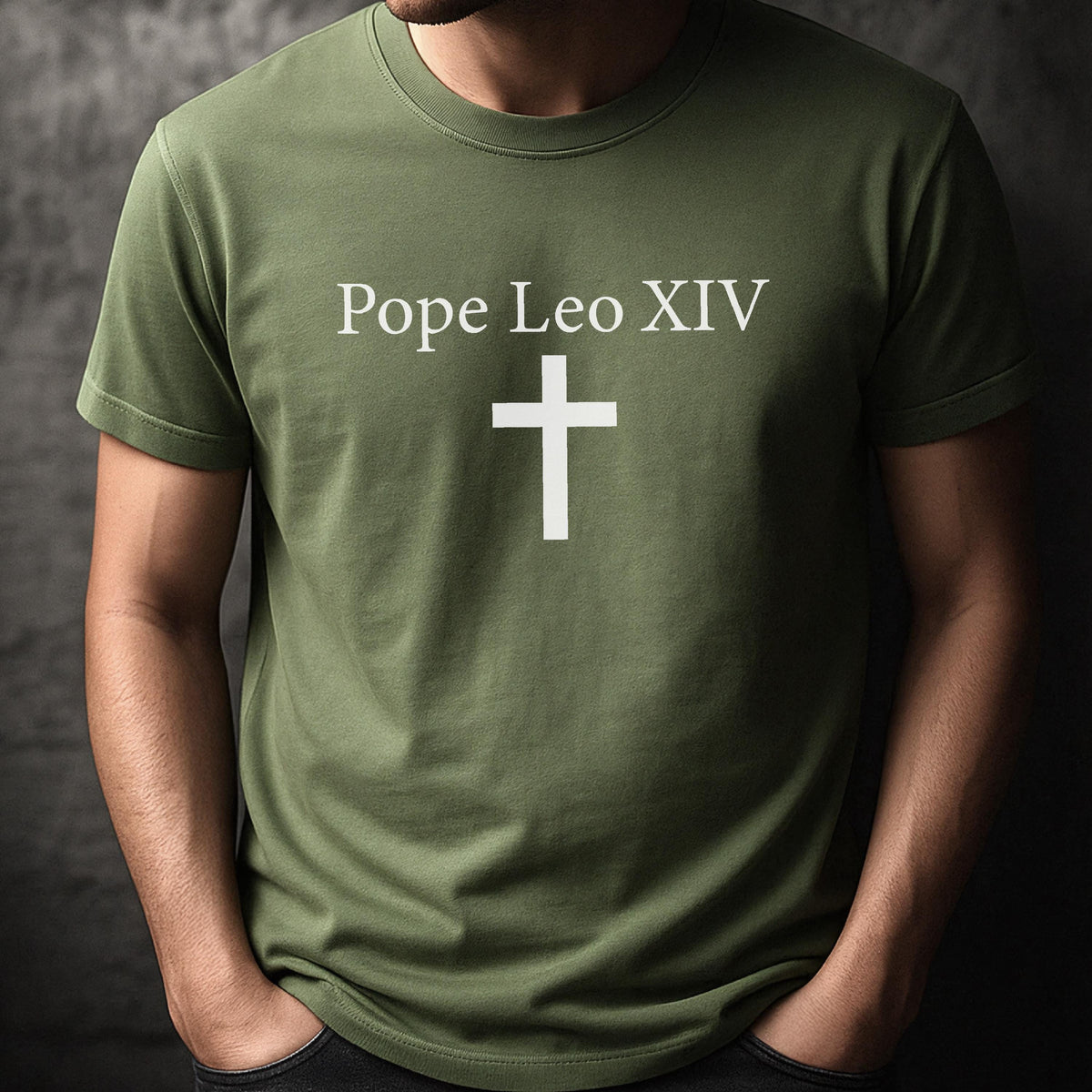 May include: Olive green t-shirt with the text "Pope Leo XIV" above a white cross. The shirt is a crew neck style and appears to be made of a soft material. The person wearing the shirt has their hands in their pockets.