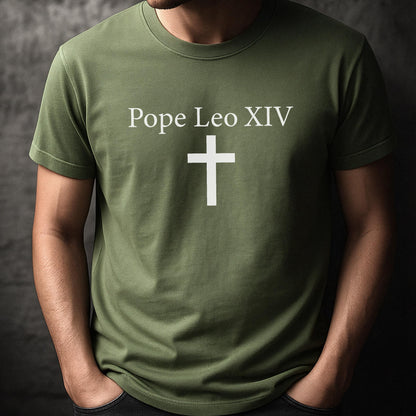 May include: Olive green t-shirt with the text "Pope Leo XIV" above a white cross. The shirt is a crew neck style and appears to be made of a soft material. The person wearing the shirt has their hands in their pockets.