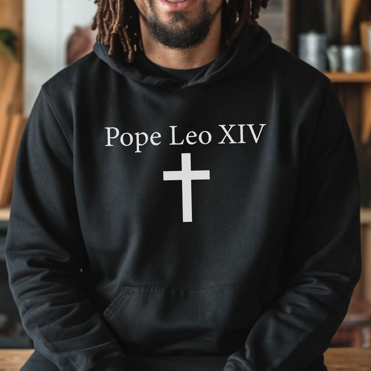May include: Black hooded sweatshirt with the text "Pope Leo XIV" above a white cross. The design is centered on the front of the sweatshirt. The sweatshirt has a front pocket and a drawstring hood.