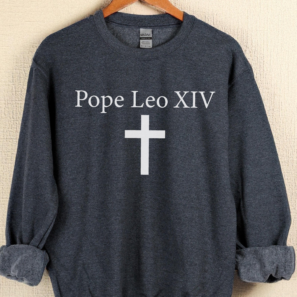 May include: Dark gray sweatshirt with the text "Pope Leo XIV" above a white cross. The sweatshirt has a crew neck and long sleeves with rolled-up cuffs. The Gildan brand label is visible at the back of the neck.