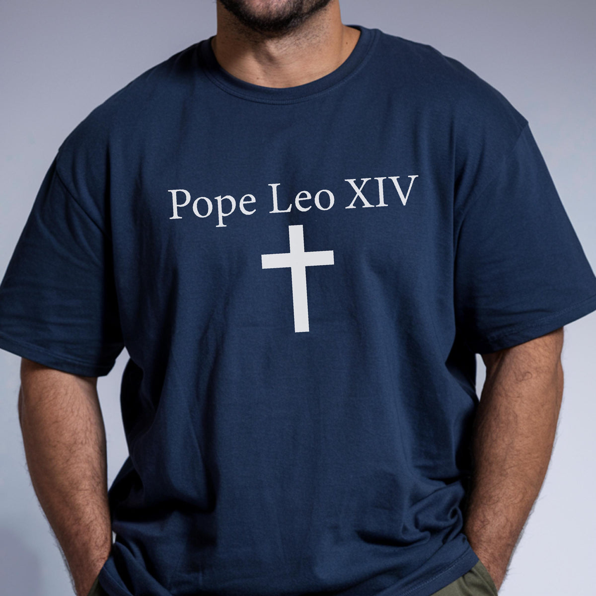 May include: Navy blue t-shirt with the text "Pope Leo XIV" above a white cross. The shirt is a crew neck style and appears to be made of a soft material. The image shows the upper body of a person wearing the shirt.