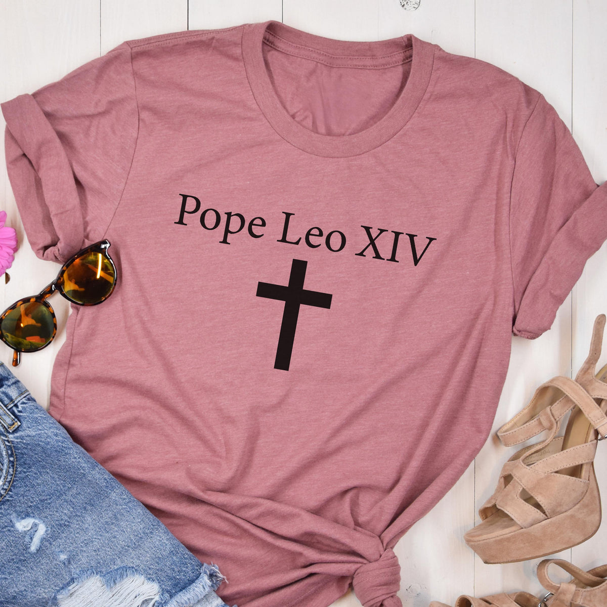 May include: A dusty rose-colored t-shirt with the text "Pope Leo XIV" above a black cross. The shirt is styled with rolled-up sleeves. Sunglasses and sandals are visible in the image.