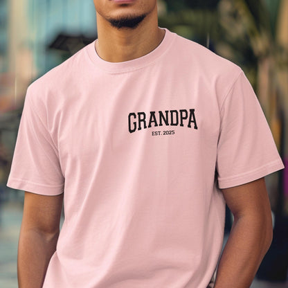 May include: A light pink t-shirt with the word "GRANDPA" printed in black, arched across the chest. Below the word "GRANDPA" is the text "EST. 2025". The shirt is a crew neck style.
