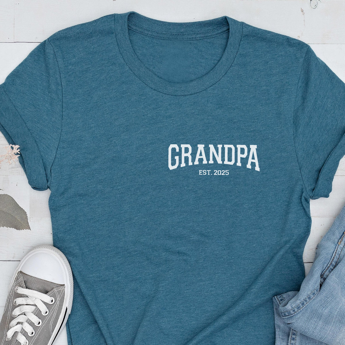 May include: Teal t-shirt with the word "GRANDPA" printed in white, with "EST. 2025" below it. The shirt is folded, with a gray sneaker and denim jeans visible in the image.
