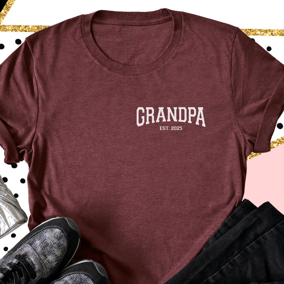 May include: A maroon t-shirt with the word "GRANDPA" printed in white, along with "EST. 2025". The shirt is made of a soft material and has a crew neck. The shirt is folded, with a pair of sneakers and black pants in the background.