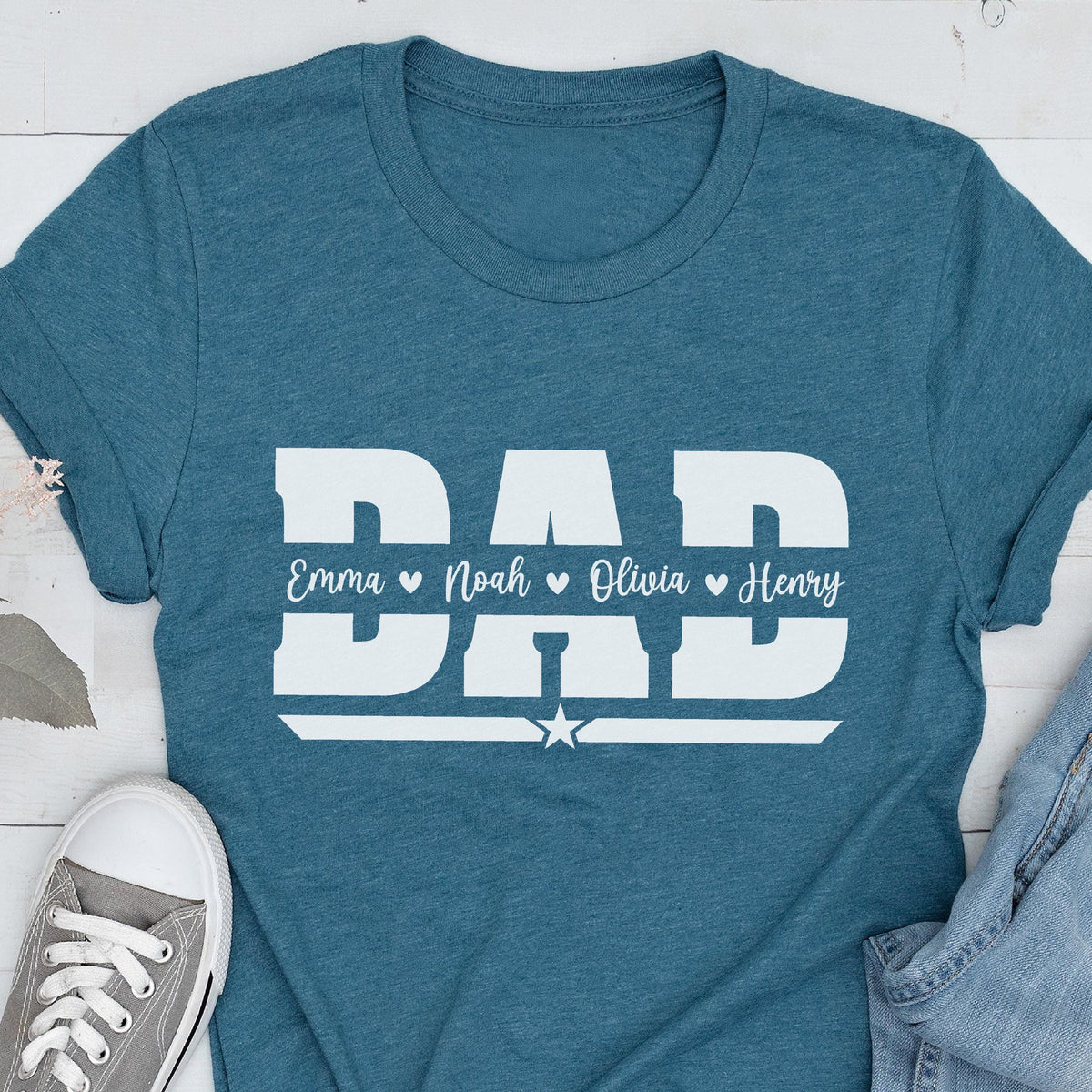 May include: Teal t-shirt with the word "DAD" in large white block letters. Below the word "DAD" are the names Emma, Noah, Olivia, and Henry. A white star is below the names. A gray sneaker and blue jeans are in the image.