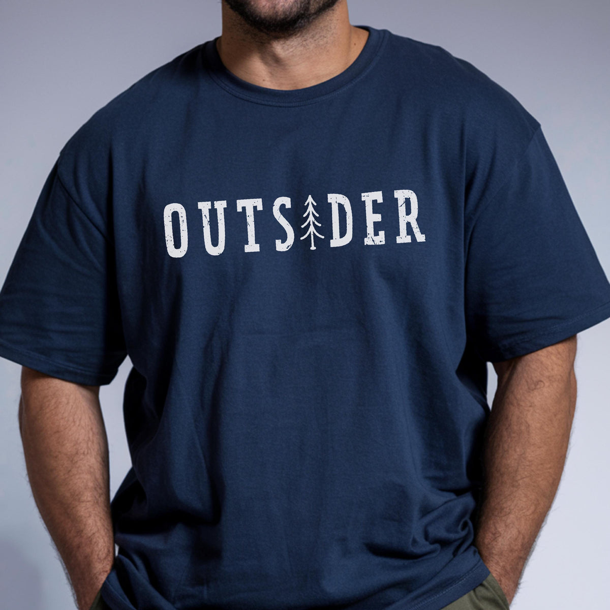 May include: A navy blue short-sleeved t-shirt featuring the white distressed text "OUTSIDER" printed across the chest. The letter "I" is replaced by a stylized graphic of a pine tree. The design has a weathered, vintage-inspired appearance.