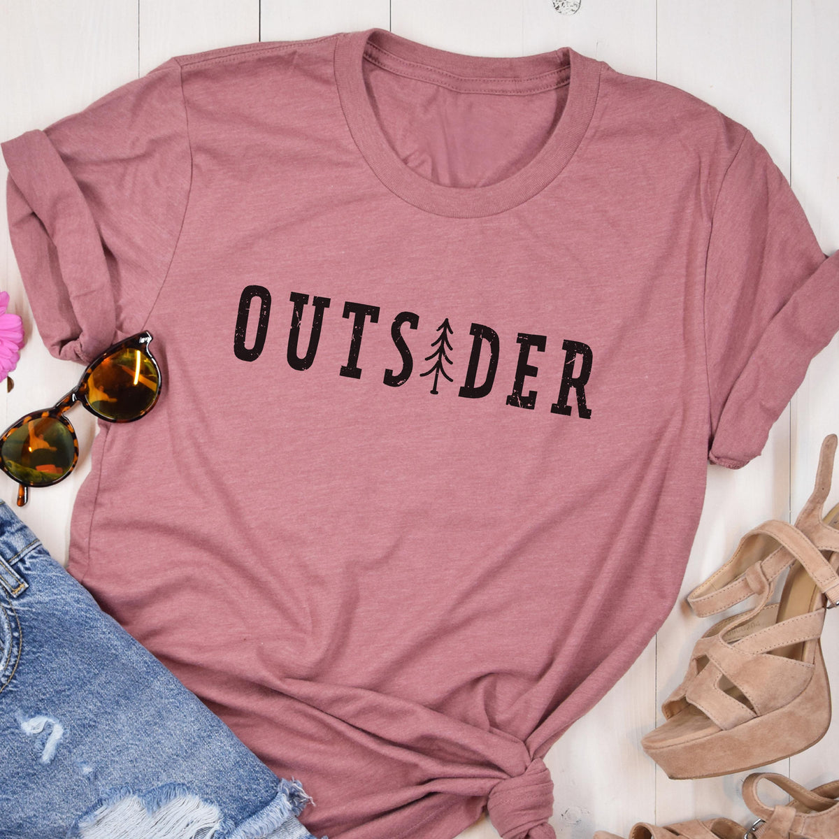 May include: A dusty rose short-sleeved t-shirt featuring the black text OUTSIDER printed across the chest, with a small stylized pine tree graphic replacing the letter I. The shirt has a crew neckline and a soft, heathered fabric texture.