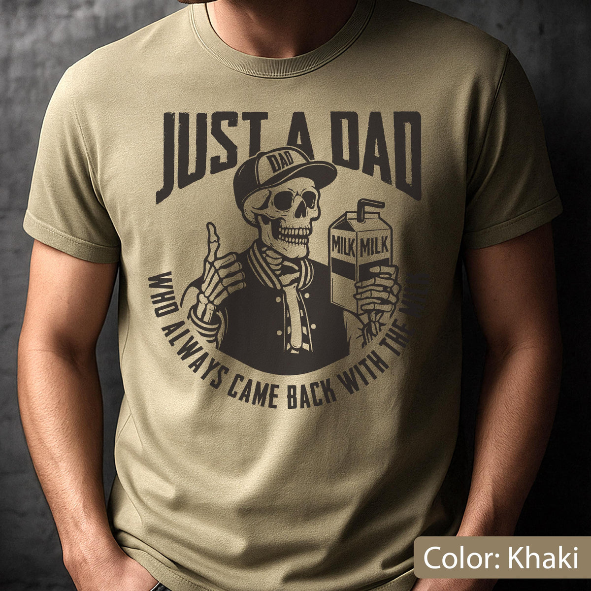 May include: Khaki-colored t-shirt with a graphic featuring a skeleton wearing a baseball cap and jacket, holding a milk carton. The text reads "JUST A DAD" and "WHO ALWAYS CAME BACK WITH THE MILK".