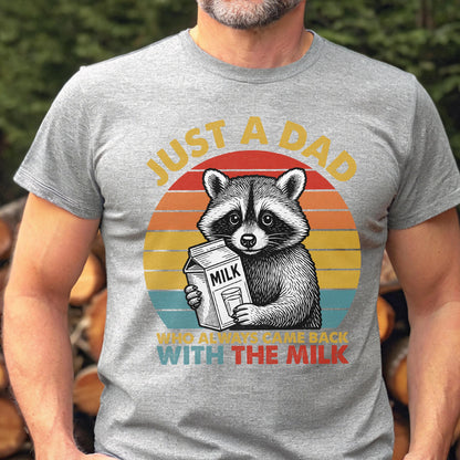 May include: A gray t-shirt features a raccoon holding a milk carton, set against a sunset design. Text reads "JUST A DAD WHO ALWAYS CAME BACK WITH THE MILK". The graphic is in shades of red, yellow, blue, and white.