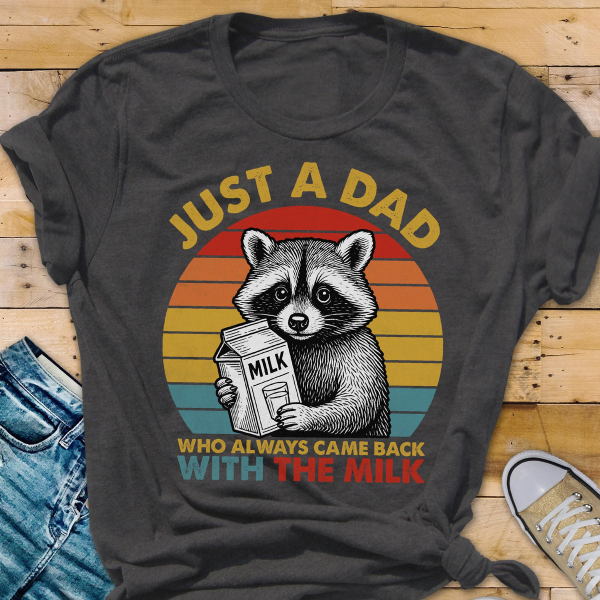 May include: Dark gray t-shirt featuring a raccoon holding a milk carton, set against a retro sunset design. Text reads "JUST A DAD" above and "WHO ALWAYS CAME BACK WITH THE MILK" below. A fun, family-themed graphic tee.