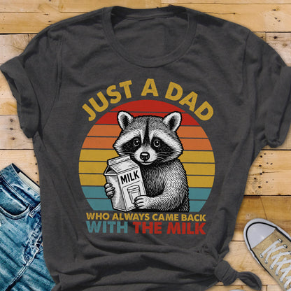 May include: Dark gray t-shirt featuring a raccoon holding a milk carton, set against a retro sunset design. Text reads "JUST A DAD" above and "WHO ALWAYS CAME BACK WITH THE MILK" below. A fun, family-themed graphic tee.