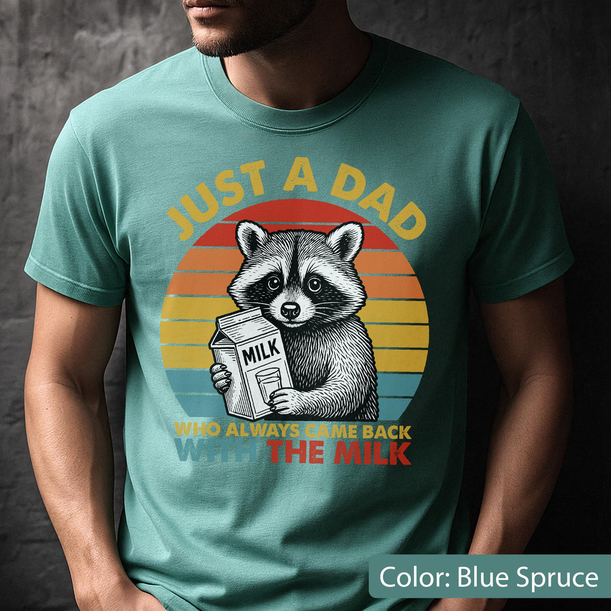May include: Blue spruce t-shirt featuring a raccoon holding a milk carton. The shirt has the text "JUST A DAD" above the raccoon and "WHO ALWAYS BRINGS ME BACK WITH THE MILK" below. The design includes a retro sunset.