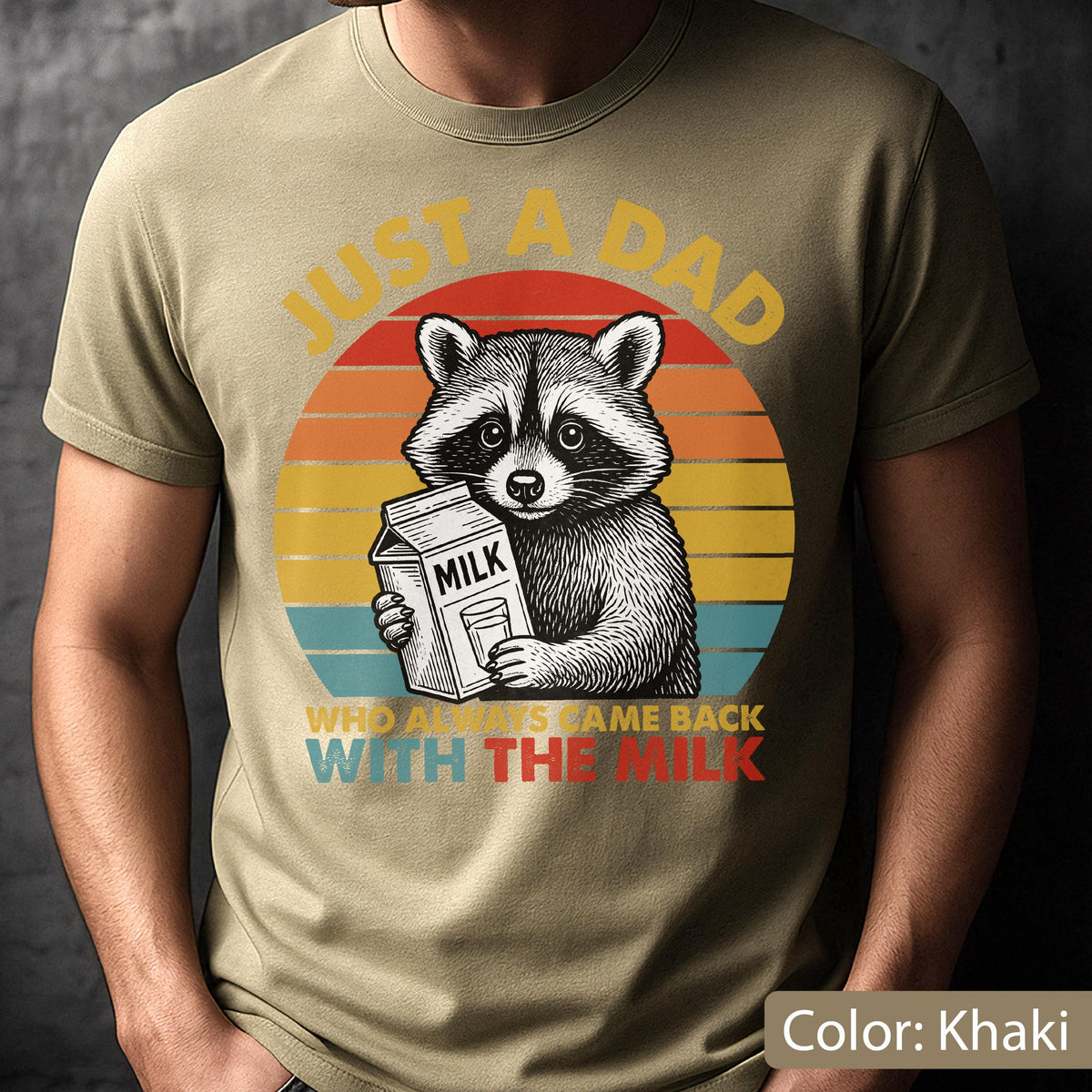 May include: Khaki-colored t-shirt featuring a raccoon holding a milk carton against a sunset design. Text reads "JUST A DAD WHO ALWAYS CAME BACK WITH THE MILK". The design is printed on a short-sleeved shirt.