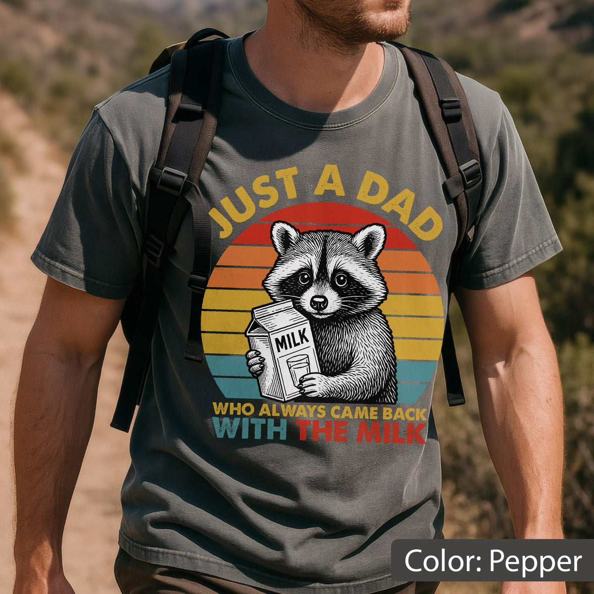 May include: Pepper-colored t-shirt featuring a raccoon holding a milk carton against a sunset backdrop. Text reads "JUST A DAD WHO ALWAYS CAME BACK WITH THE MILK" in yellow and red. The shirt is a casual wear item.