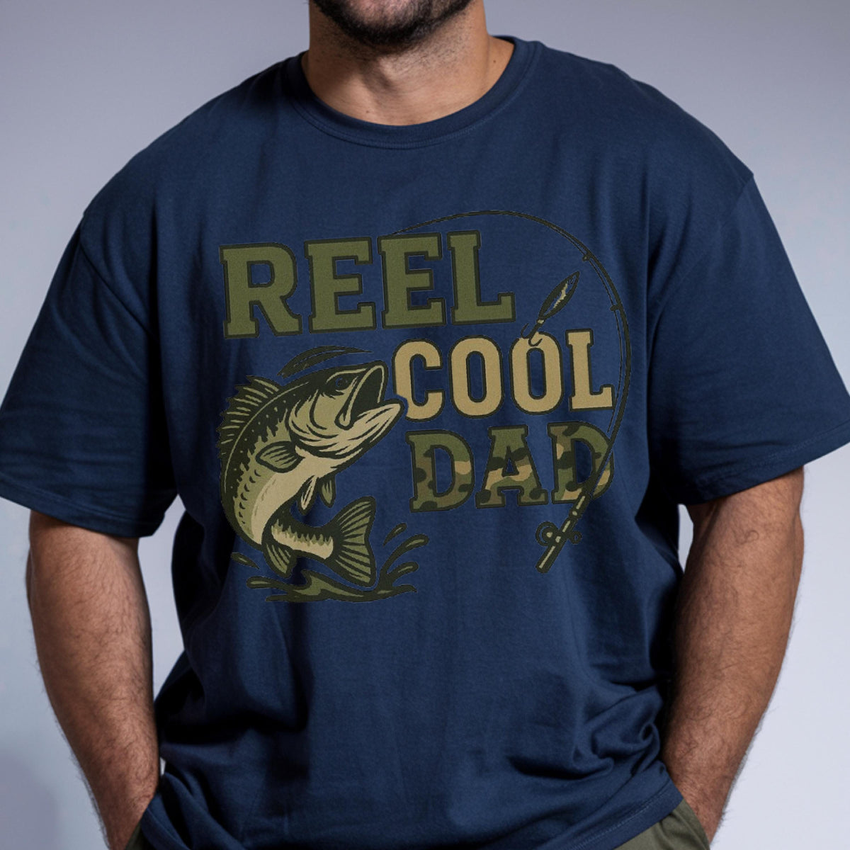 May include: A navy blue short-sleeve t-shirt featuring a graphic print of a fish jumping out of water with a fishing rod and line. The text REEL COOL DAD is printed in olive green and camouflage patterns across the center of the chest.