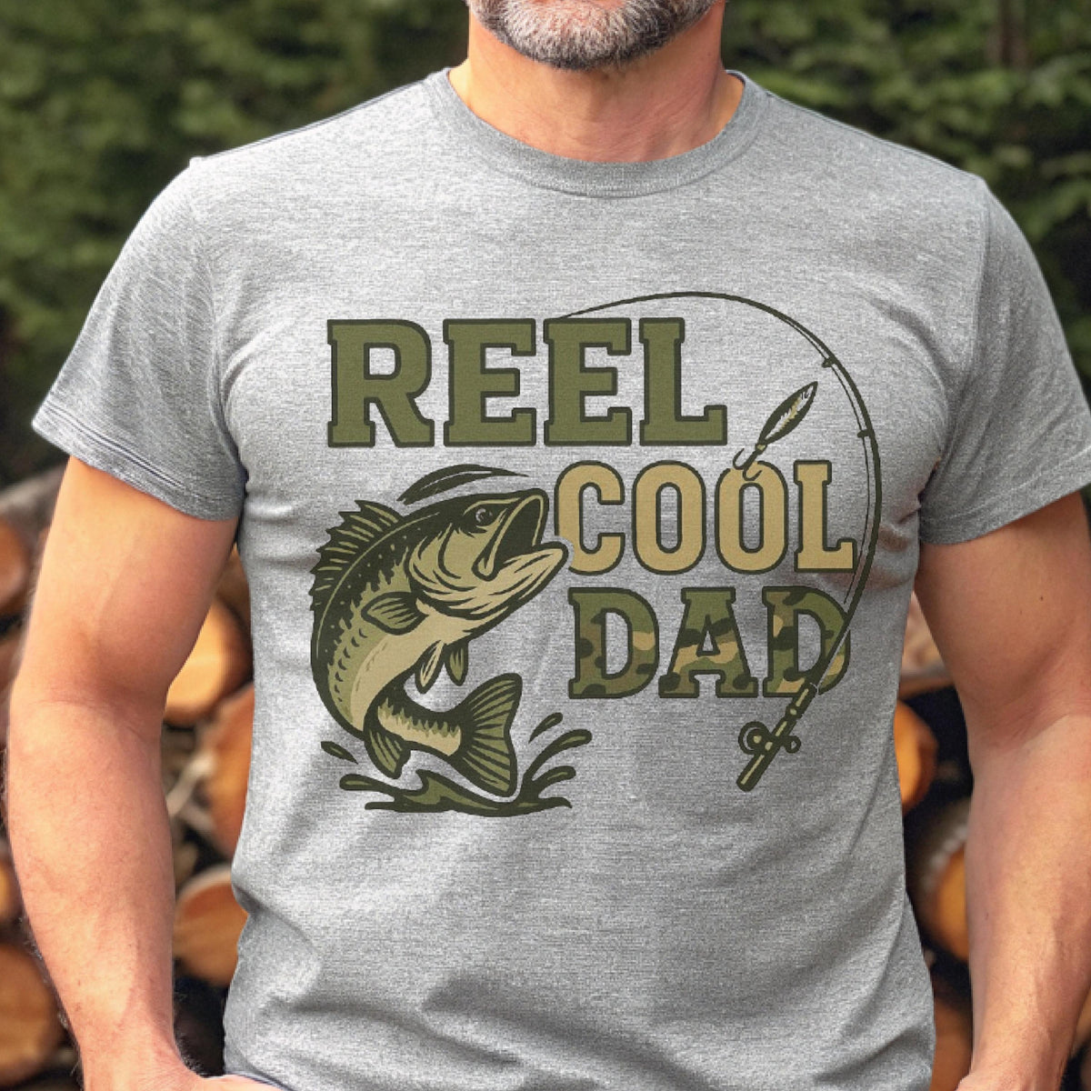 May include: A heather gray short-sleeve t-shirt featuring a graphic of a jumping fish and a fishing rod. The text REEL COOL DAD is printed in a camouflage pattern. The design includes a curved fishing line extending from the rod to the fish.